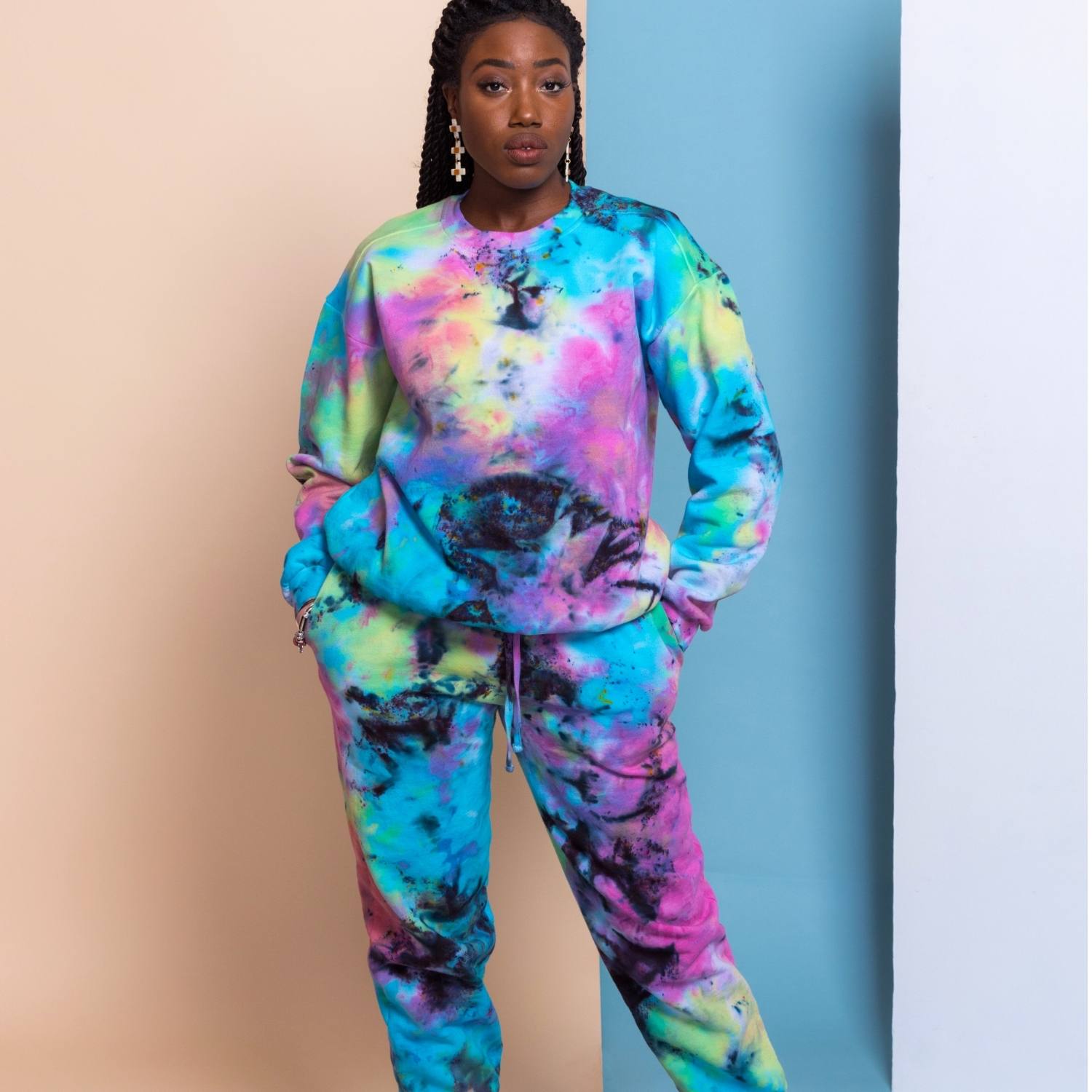 http://mashaapparel.com/cdn/shop/files/Woman-standing-in-bright-tie-dye-sweatsuit-hands-in-pockets-two-tone-blue-pink-background.jpg?v=1706038949